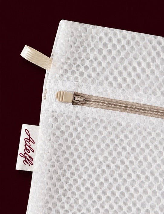 A close-up of a white mesh zippered pouch with a repeating circular pattern, featuring a cream-colored pull tab and a small fabric tag with the brand name 'Adoree' in cursive, set against a dark burgundy background.
